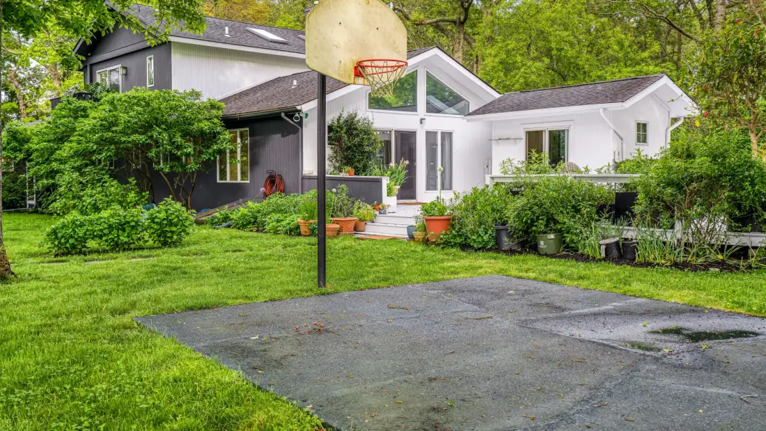 Una casa y un jardín junto a una canasta de baloncesto y una cancha pavimentada.