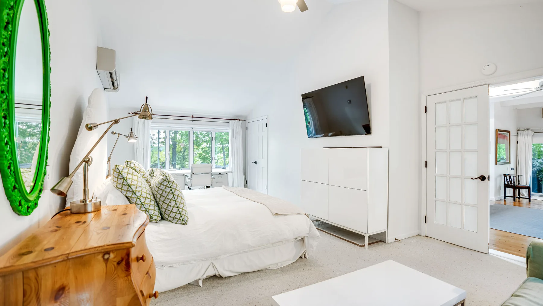 Un dormitorio luminoso con una cama, una cómoda de madera y un televisor en la pared.