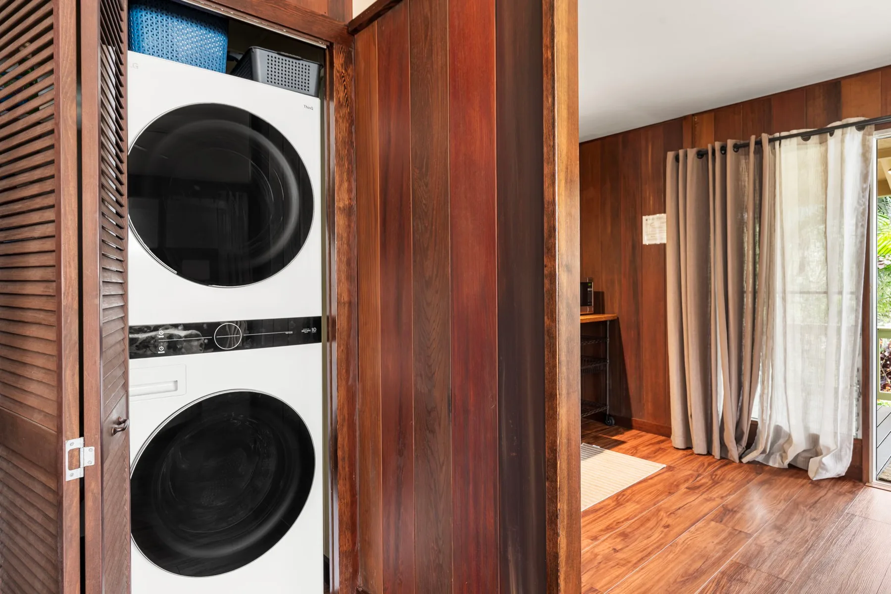 Una habitación revestida de madera con una lavadora y secadora apiladas junto a ventanas con cortinas.