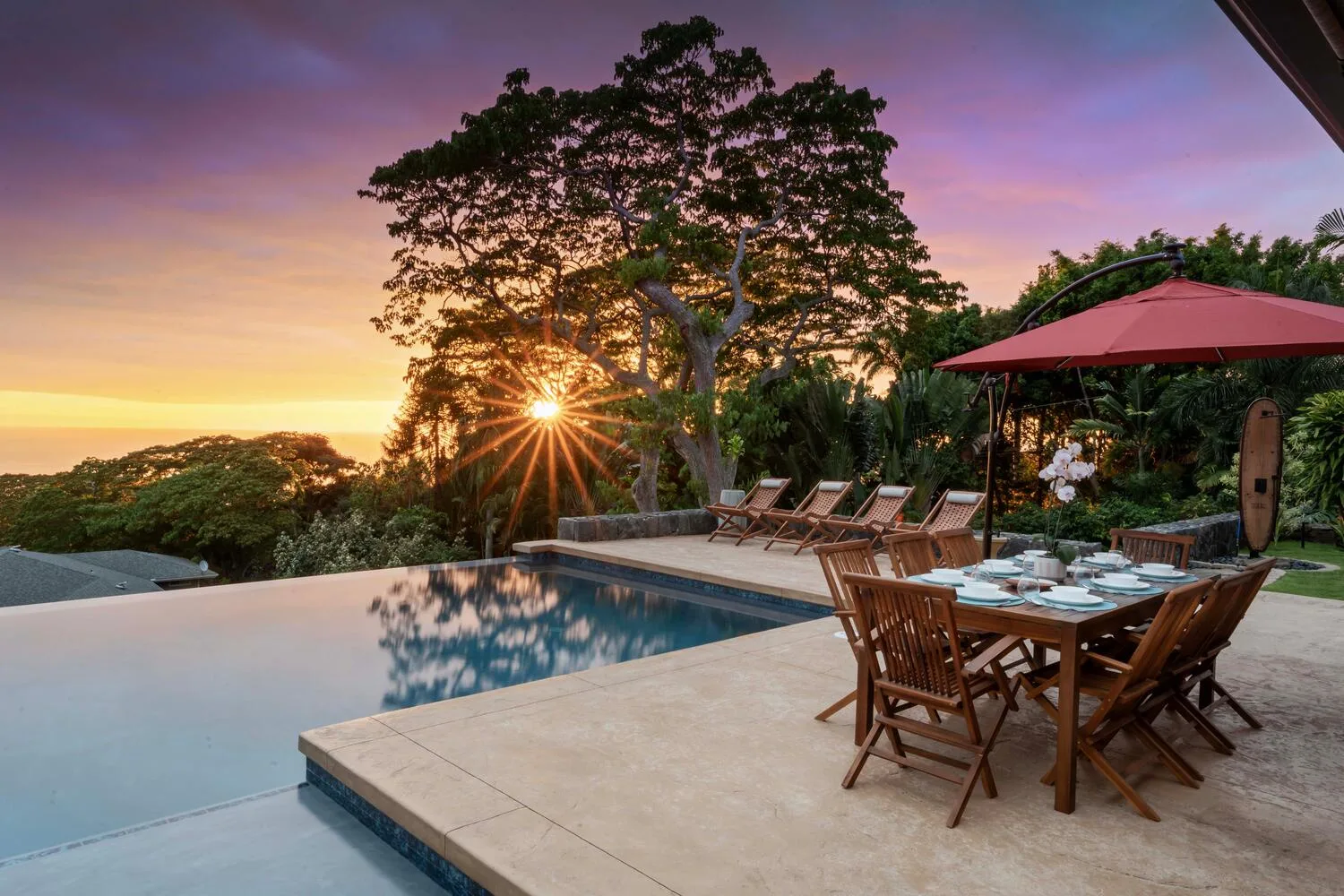 Una piscina al aire libre y una zona de comedor miran hacia los árboles bajo un cielo colorido al atardecer.