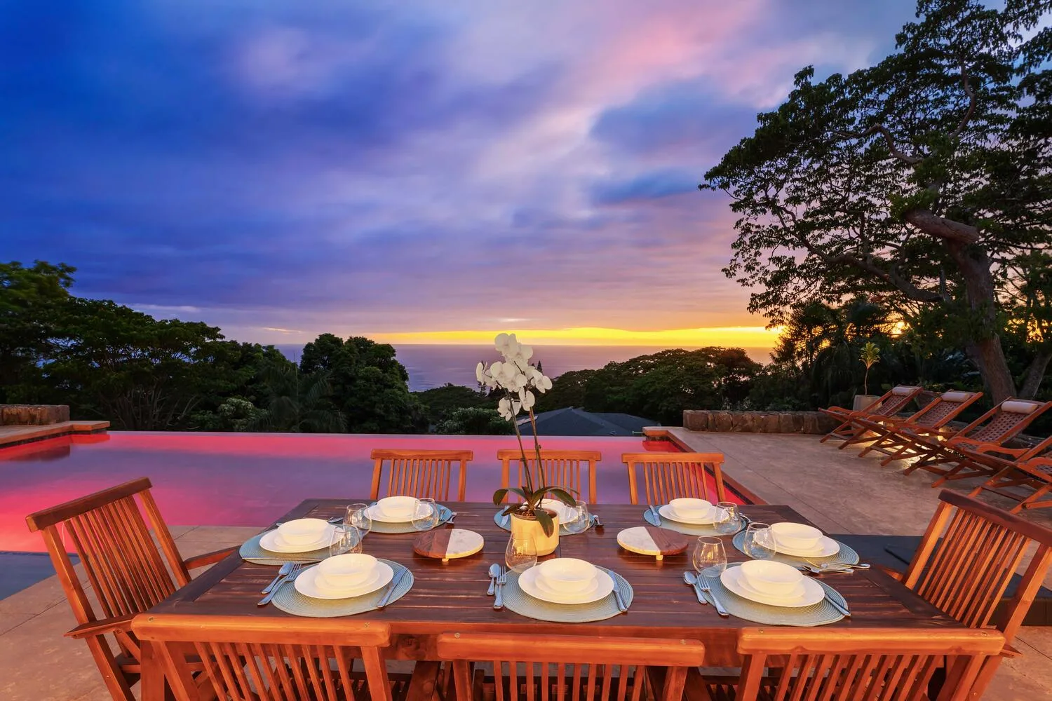 Una mesa de comedor junto a una piscina ofrece vistas de árboles y un cielo colorido al atardecer.