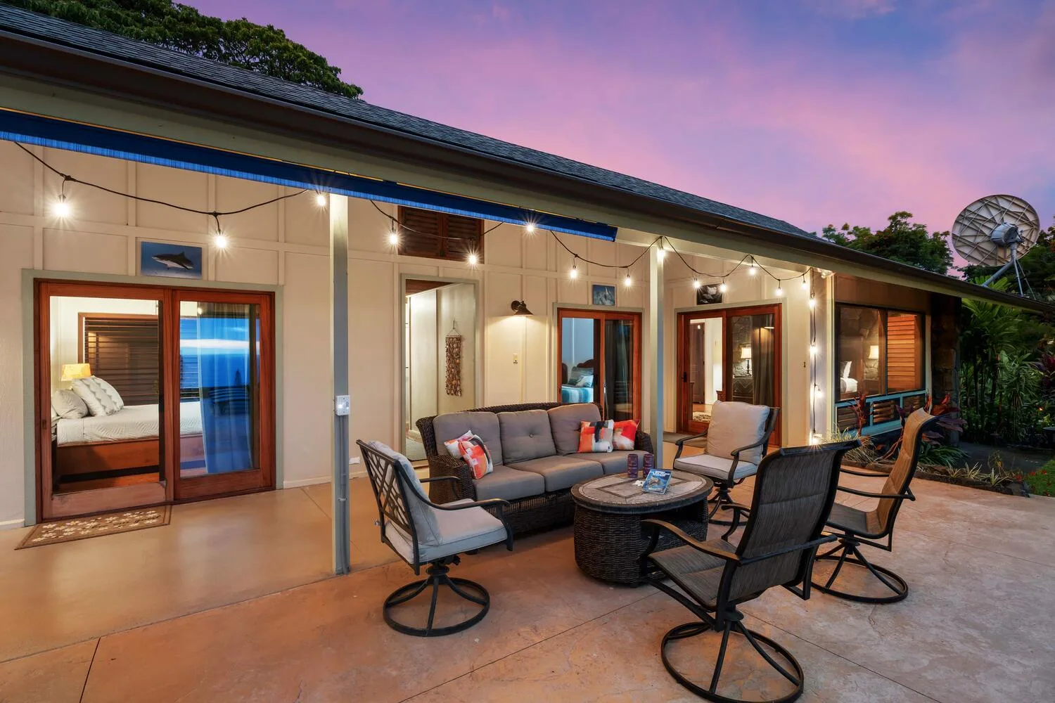 Un patio cubierto con asientos al aire libre y luces colgantes junto a la casa.
