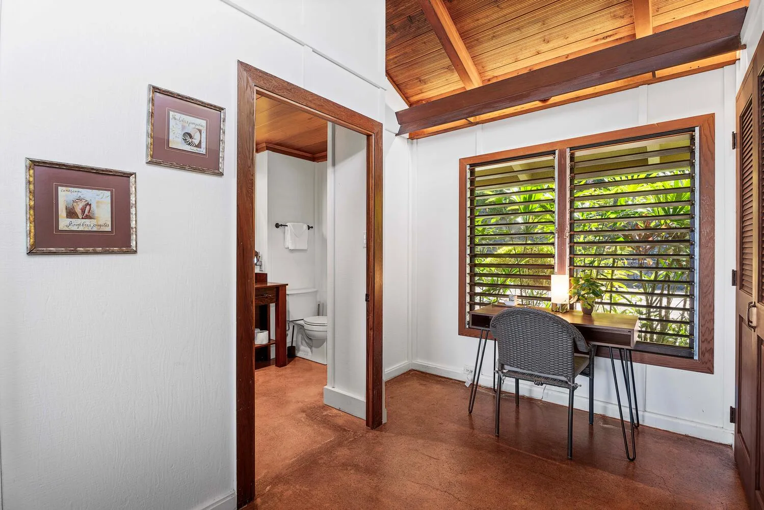 Una habitación luminosa tiene una mesa pequeña y asientos junto a amplias ventanas de celosía.