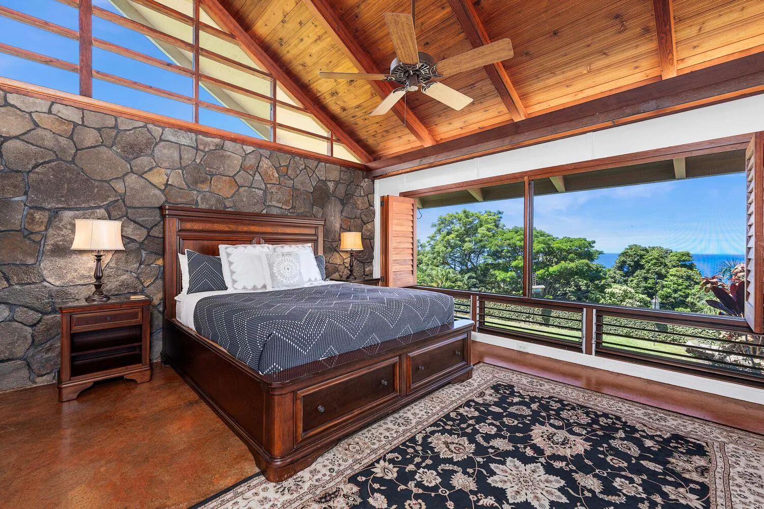 Un dormitorio con una cama grande y amplias ventanas con vistas a árboles y al océano.