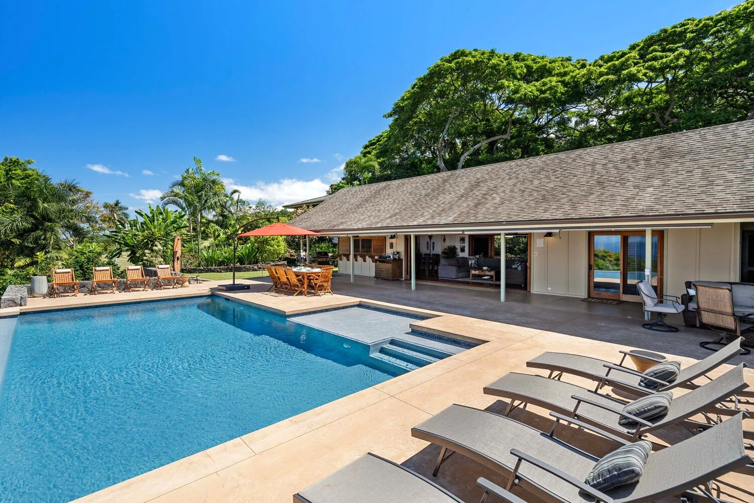 Una piscina al aire libre junto a un patio cubierto con asientos y vegetación tropical.