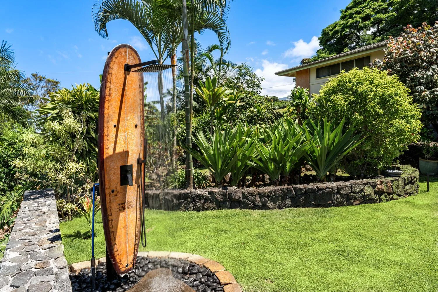 Una ducha de jardín está junto a un césped verde y plantas tropicales frondosas.