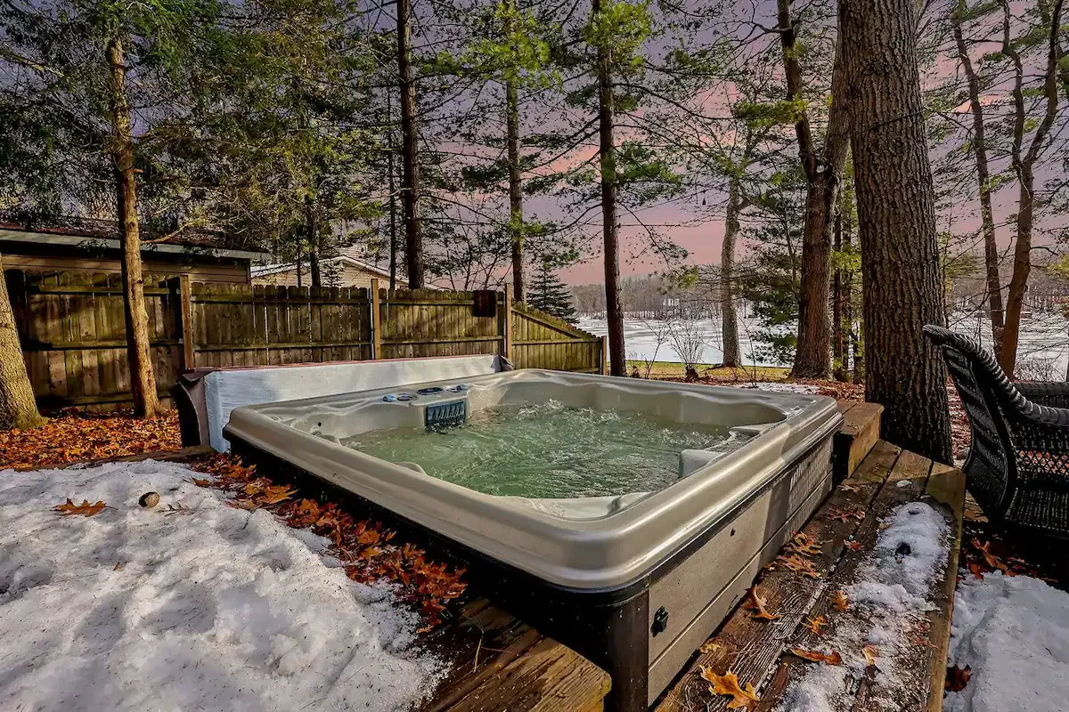 Una bañera de hidromasaje al aire libre está sobre una terraza de madera entre árboles, con nieve en el suelo.