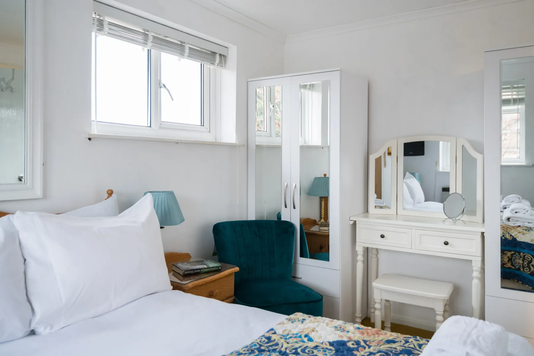 Un dormitorio luminoso con cama, asiento verde azulado y muebles con espejos.