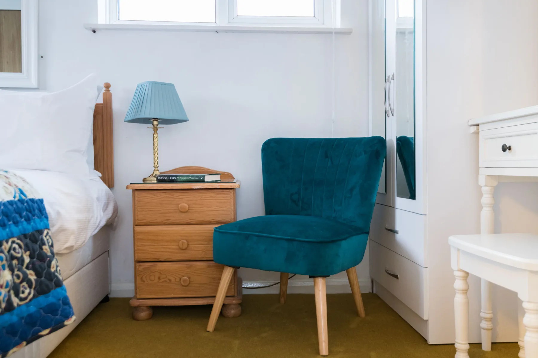 Un dormitorio con cama, mesita de madera y asiento tapizado de color verde azulado.