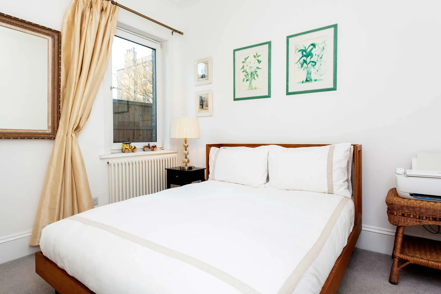 Un dormitorio luminoso con una cama blanca, cuadros enmarcados y una ventana con cortinas.