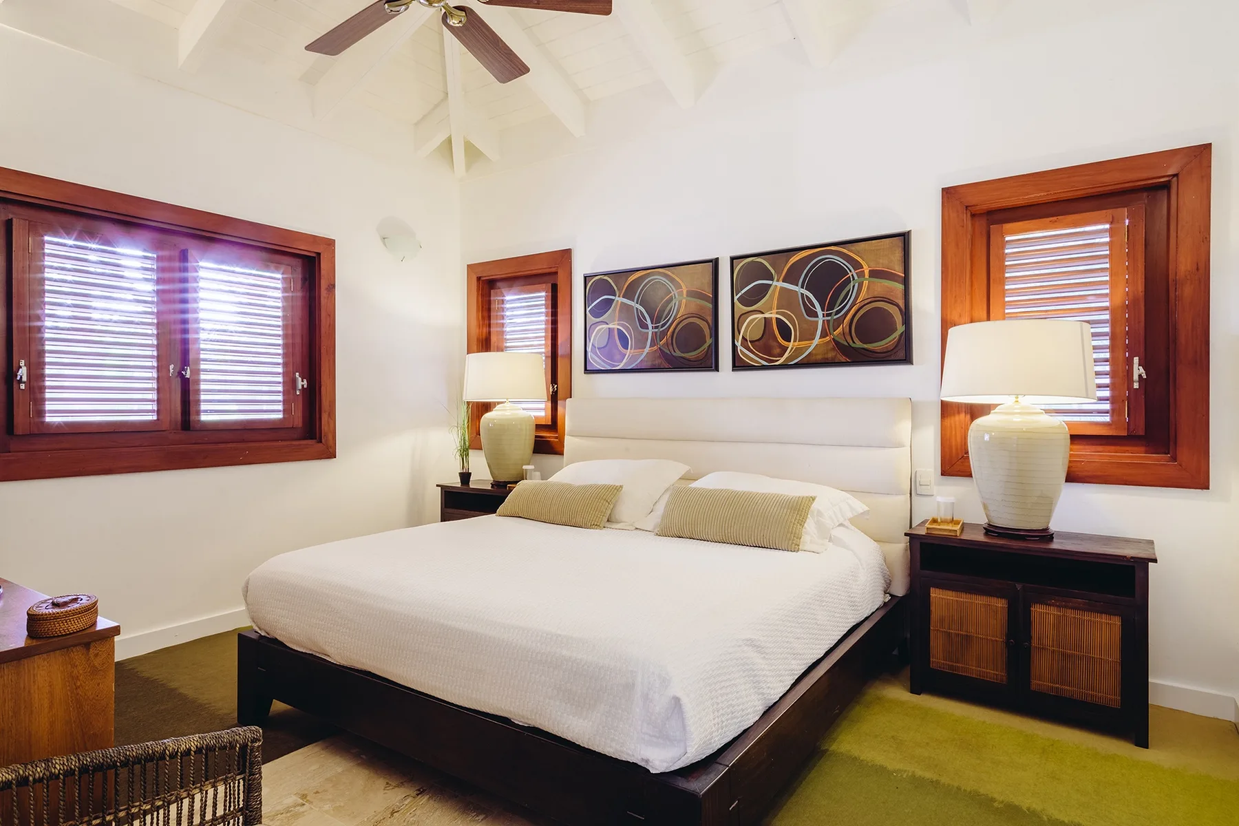 Un dormitorio con una cama bien tendida, cuadros enmarcados y ventanas con persianas de madera.