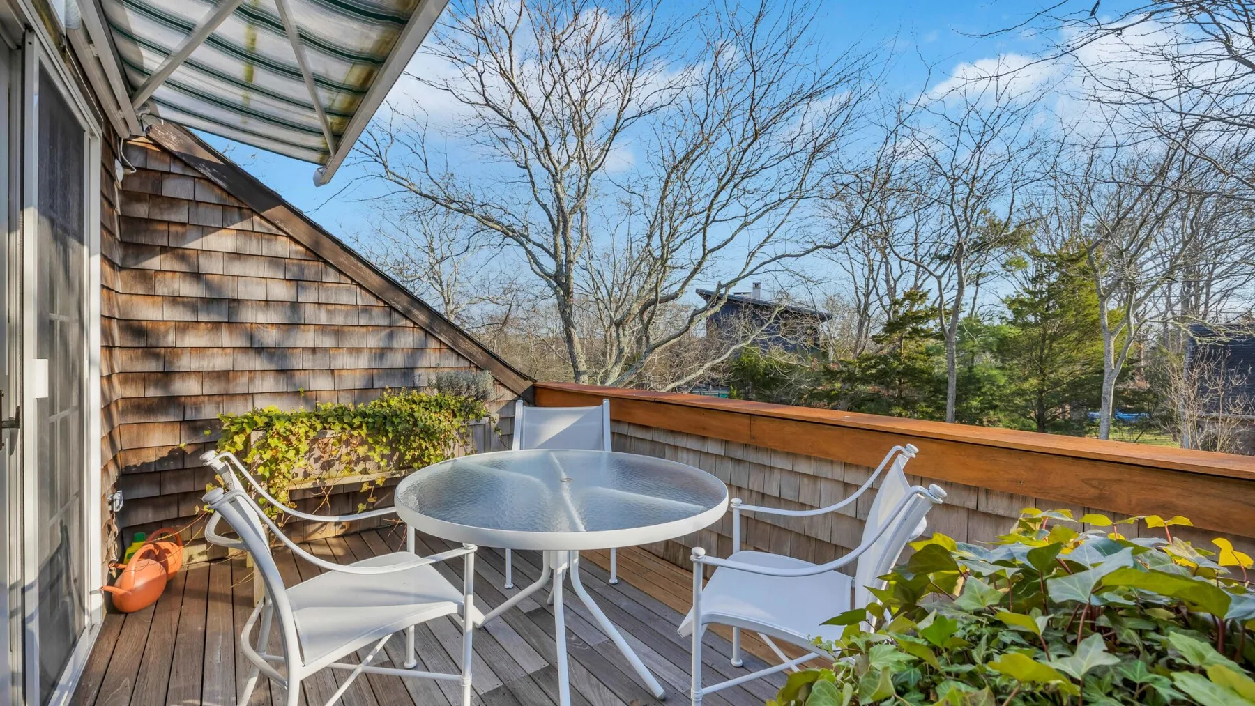 A deck with white seating and a round glass table overlooks leafy trees.