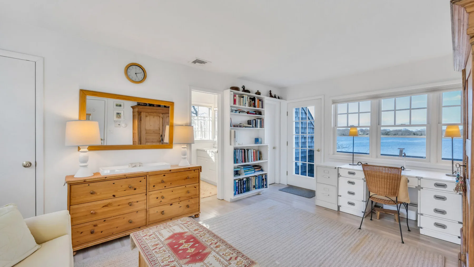A bright bedroom with a wooden dresser, bookshelves, and a desk by the windows.