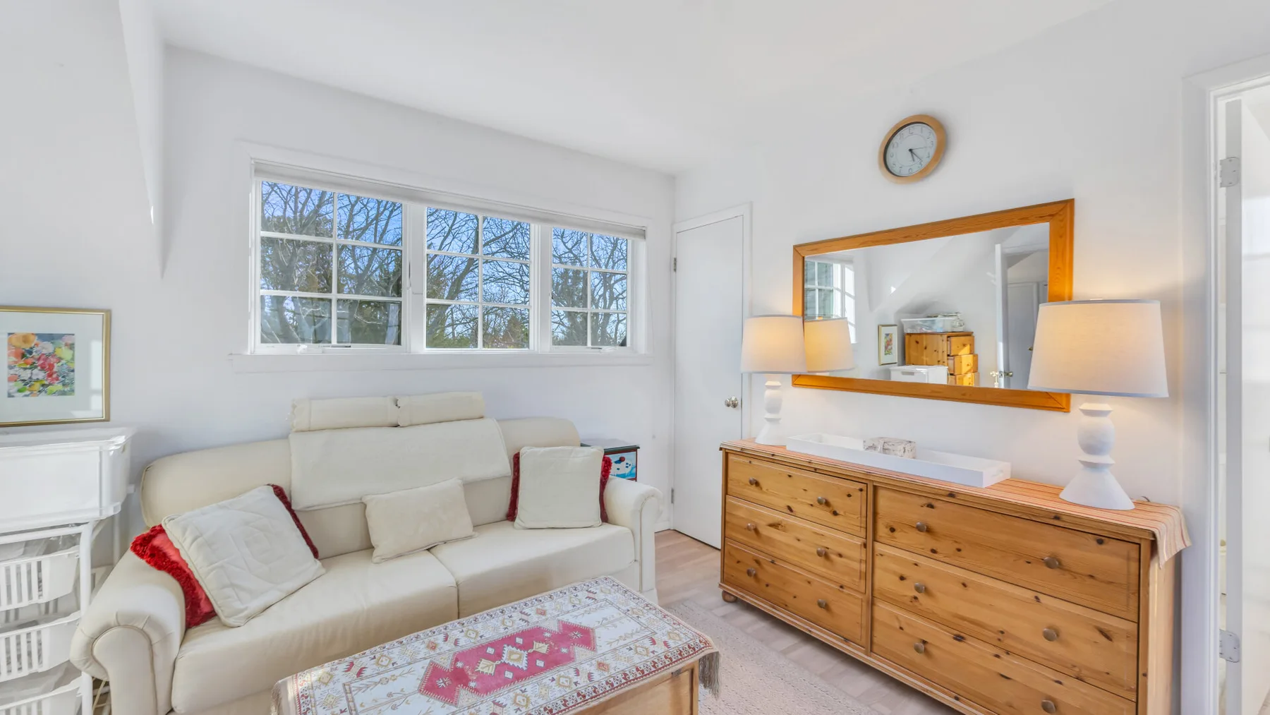 A bright living room has a cream sofa, a wooden dresser, and a large mirror.