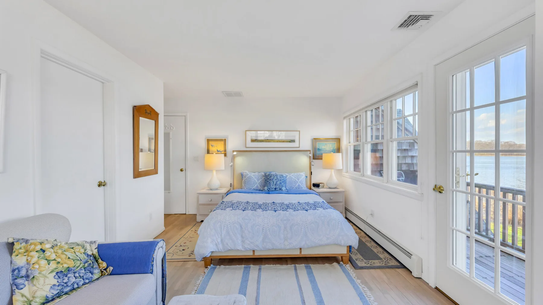 A bright bedroom has a bed between bedside tables and windows overlooking the water.