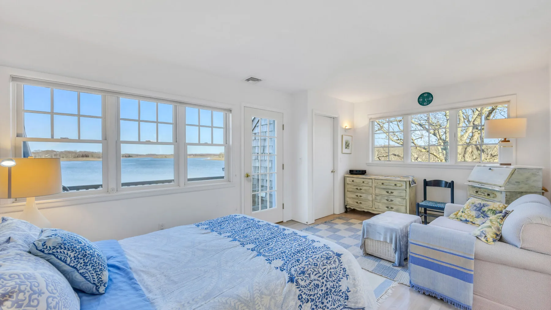 A bright bedroom with a large bed, blue patterned bedding, and windows overlooking the water.