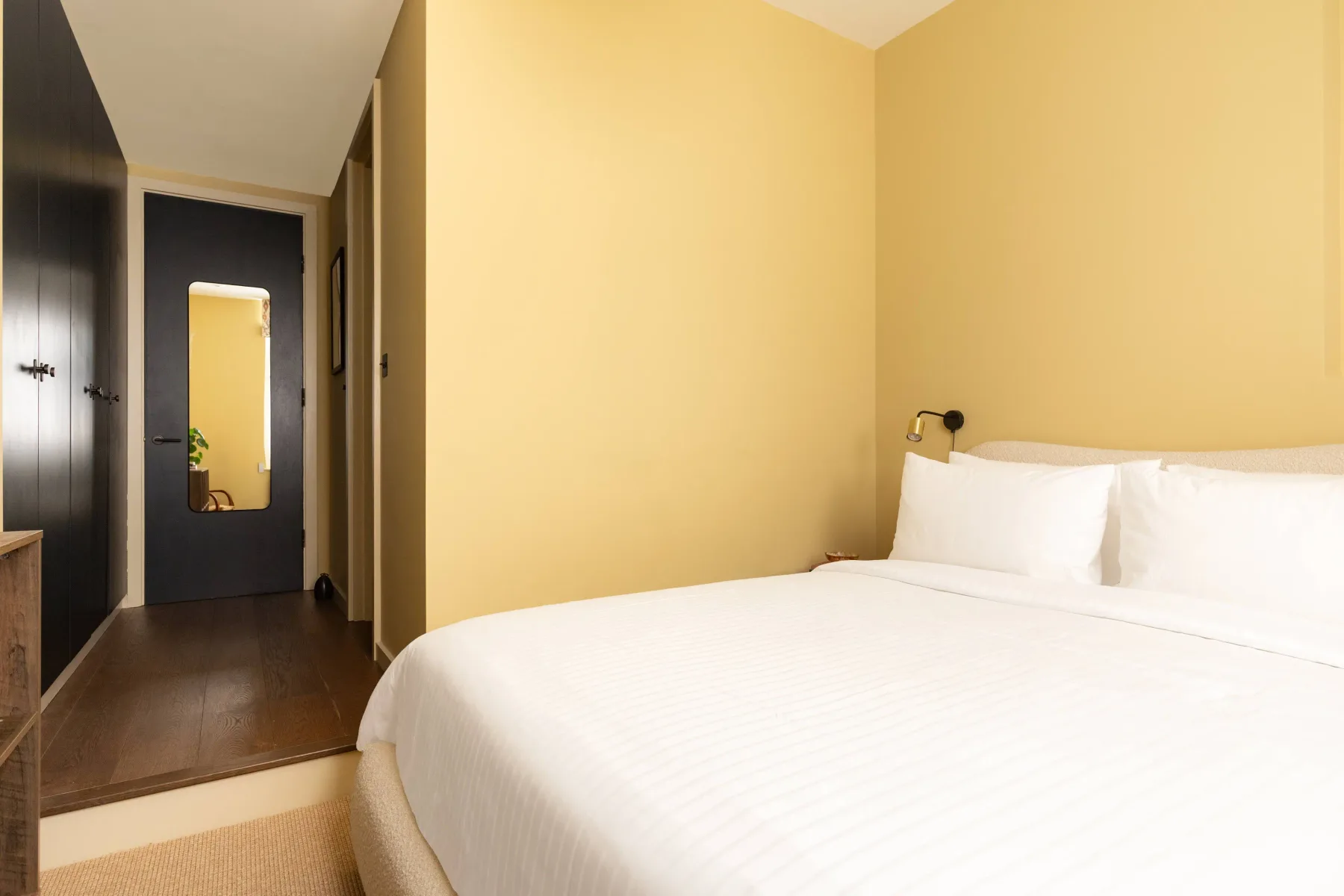 Un dormitorio con una cama blanca junto a un pasillo y una puerta oscura.