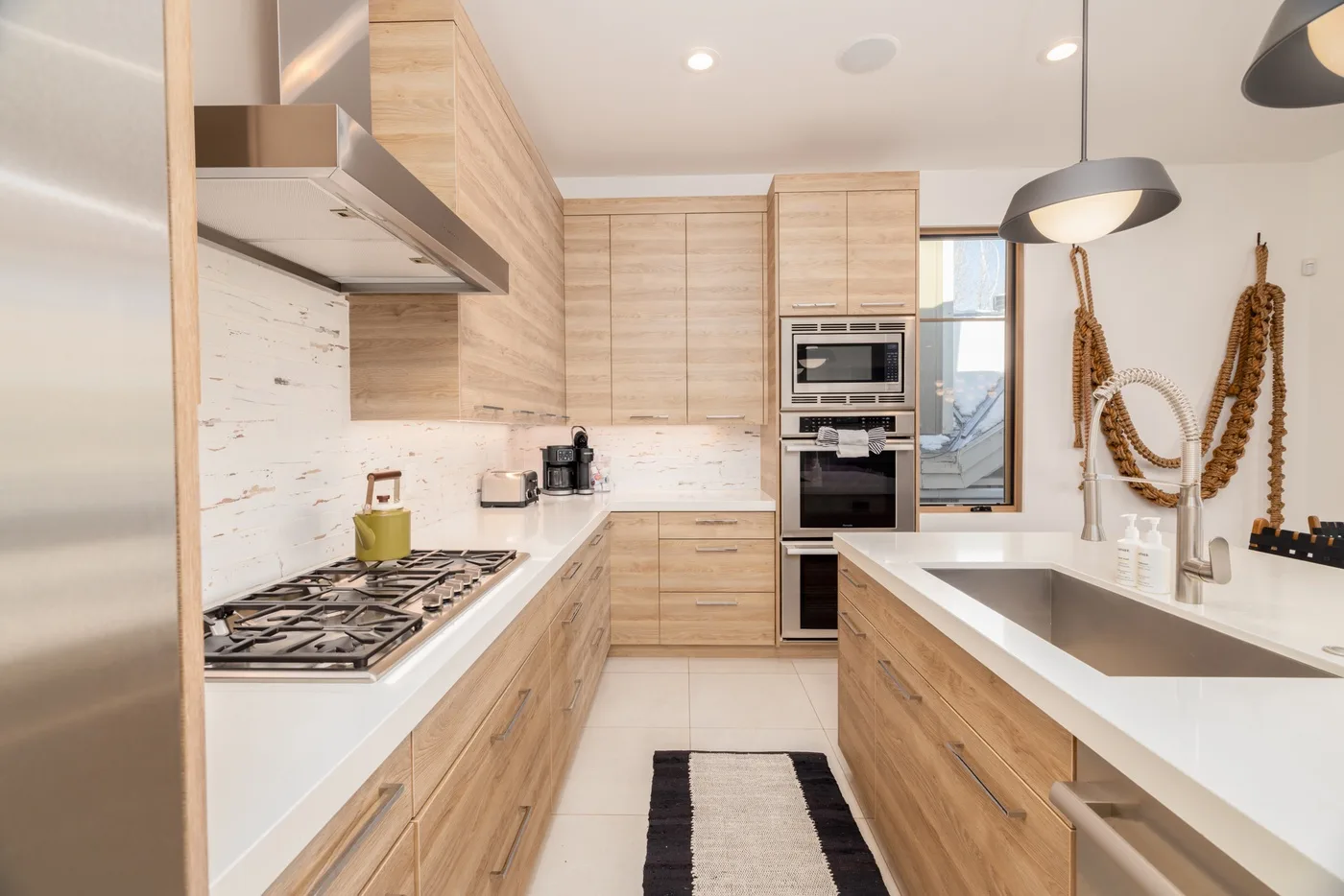 A modern kitchen with light wood cabinets, stainless steel appliances, and a central walkway.
