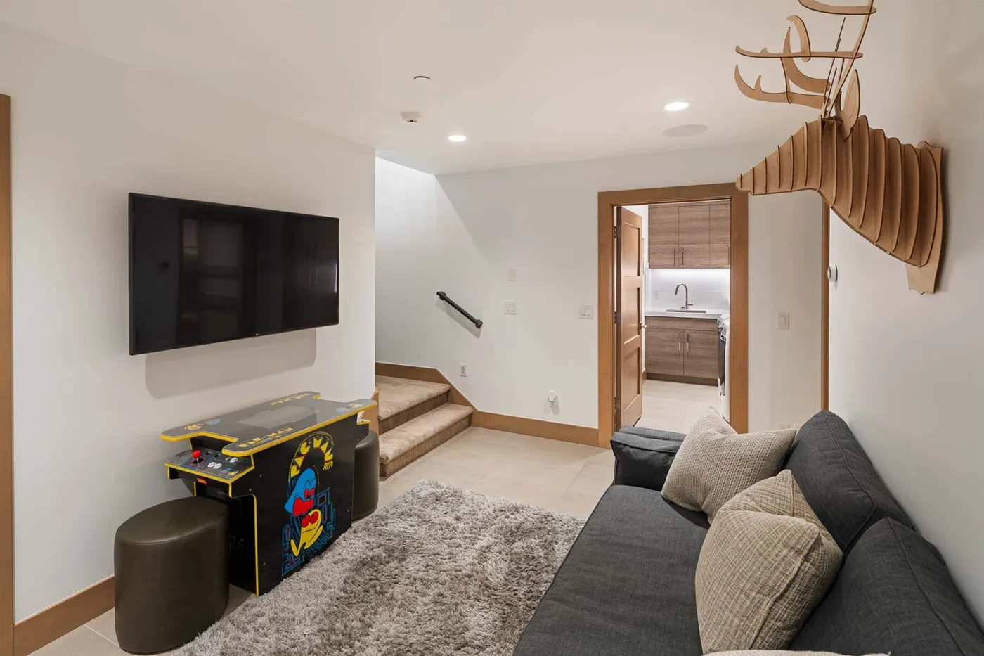 A bright living area with a dark sofa, wall-mounted television, and arcade game.