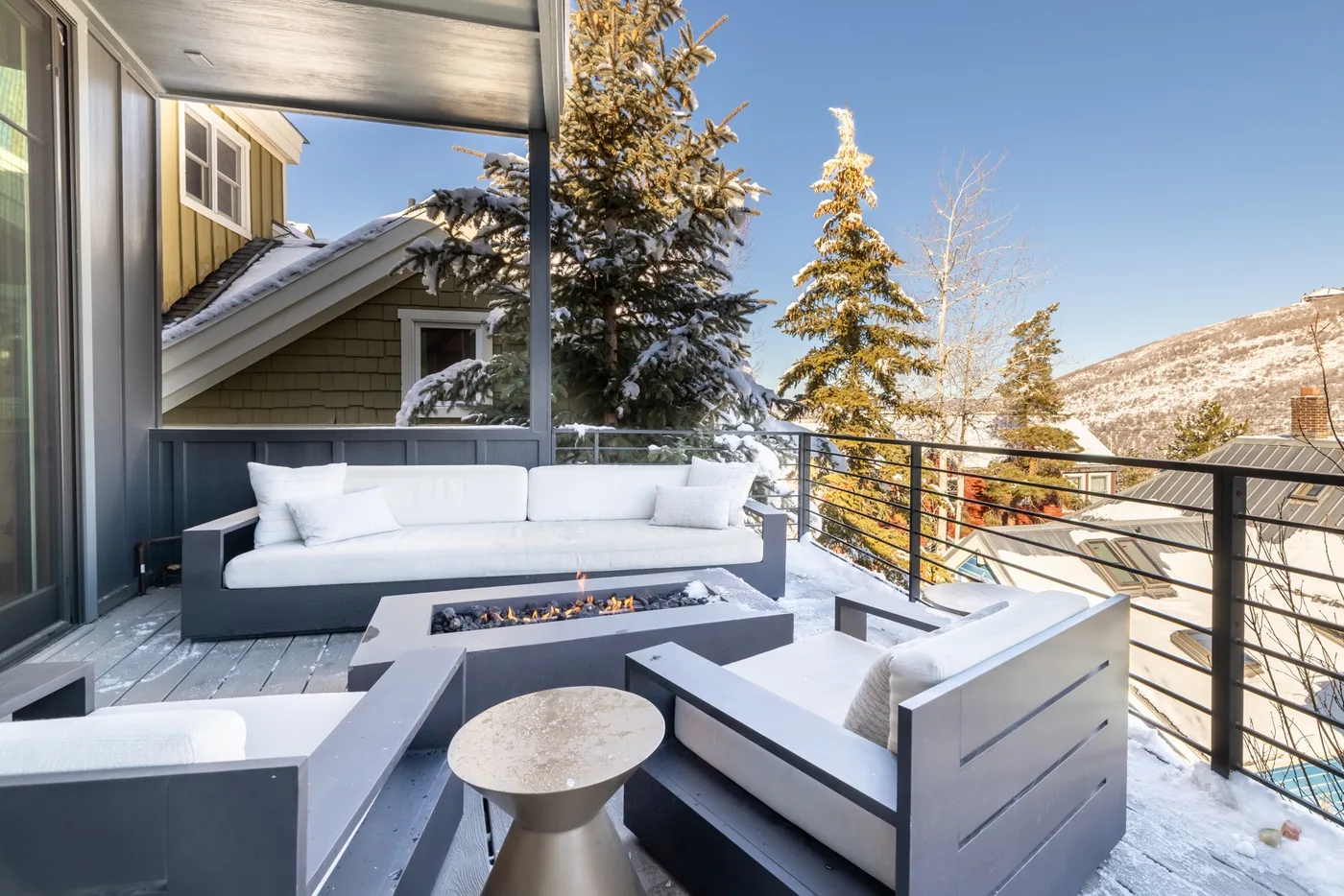 An outdoor deck seating area with a low fire table overlooks a snowy mountain landscape.