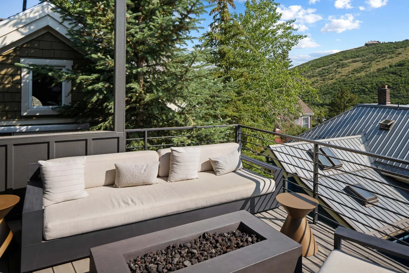 An outdoor deck with cushioned seating and a rectangular fire pit overlooks a green hillside.