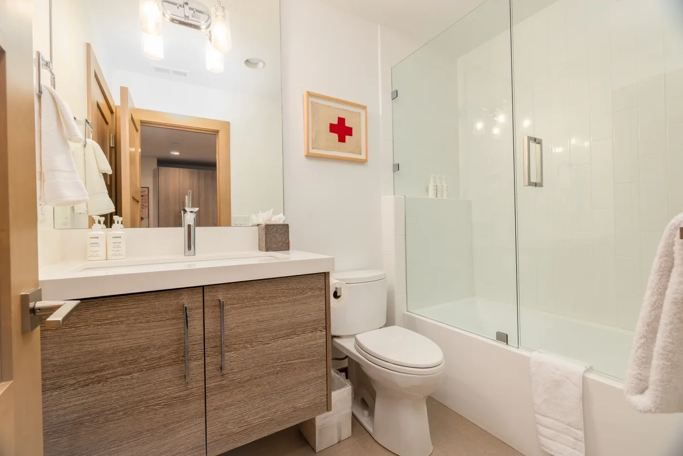 A bright bathroom with a vanity, toilet, and bathtub behind a glass screen.