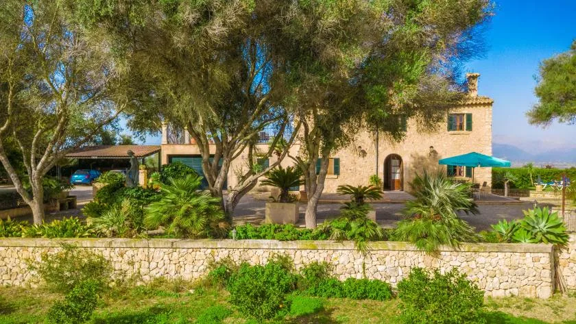 Un edificio de piedra se alza tras un muro bajo, a la sombra de árboles y rodeado de vegetación.