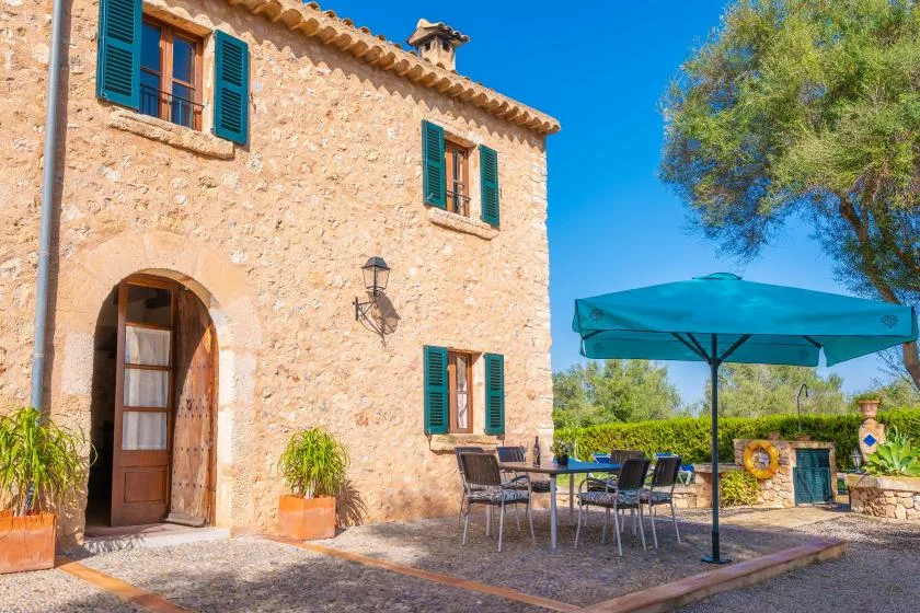 Una casa de piedra con contraventanas verdes da a un patio exterior con asientos y un toldo.