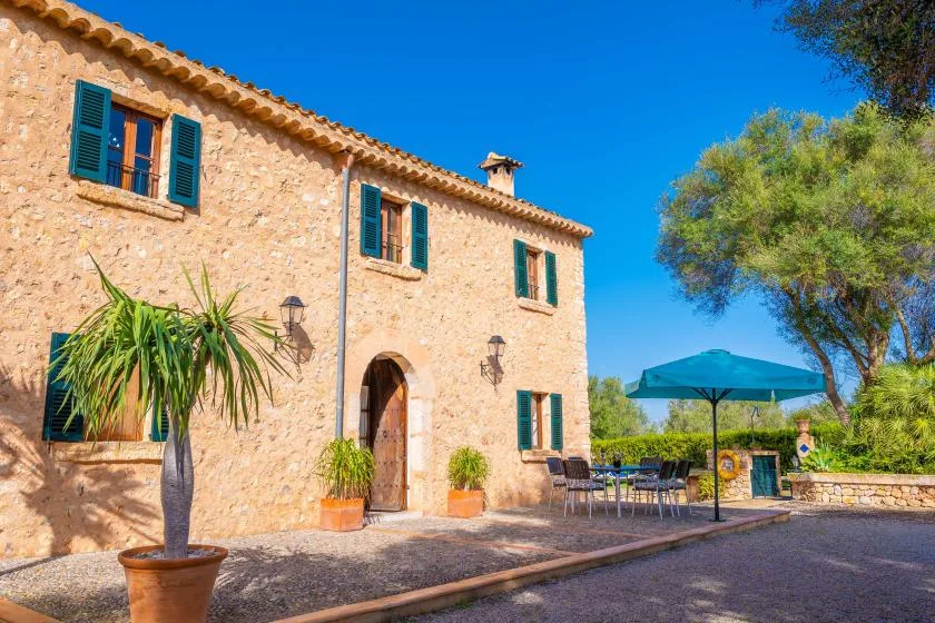 Una casa de piedra con contraventanas turquesas está junto a una zona de estar al aire libre con sombra.