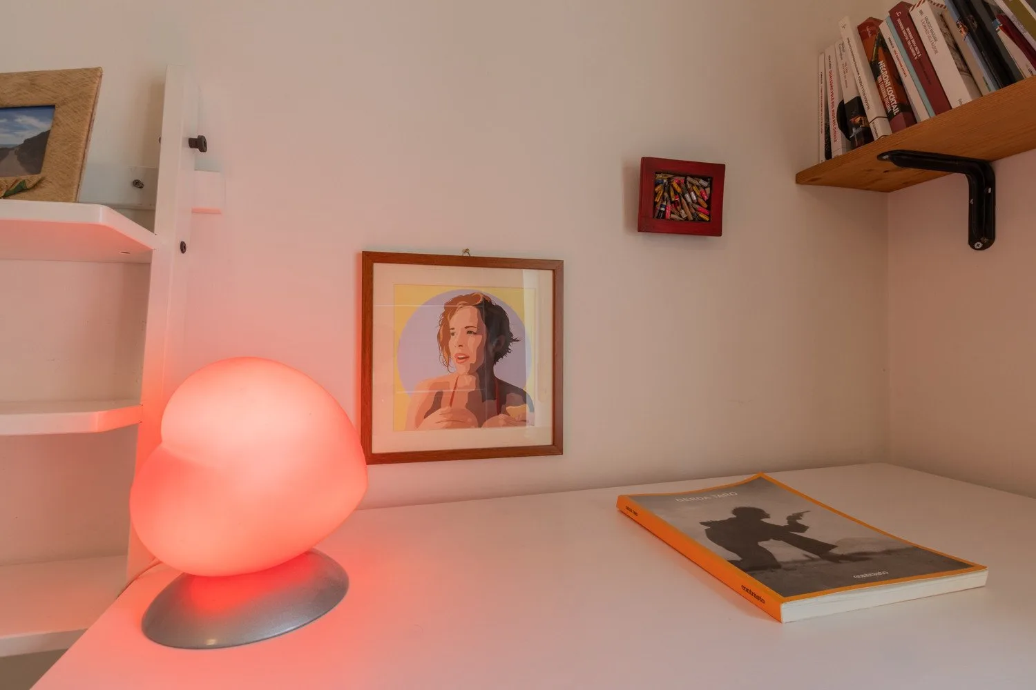 Un escritorio muestra cuadros, una lámpara encendida y un libro bajo un estante de pared.