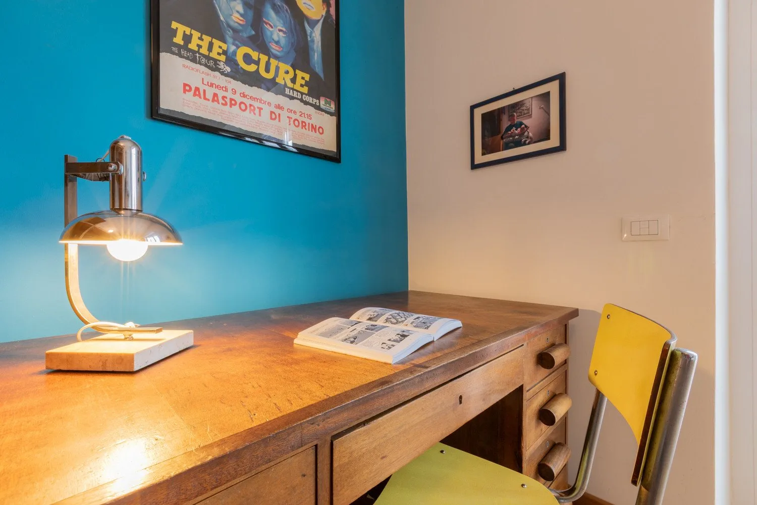 Un escritorio de madera con una lámpara encendida y un libro abierto está junto a una pared turquesa.