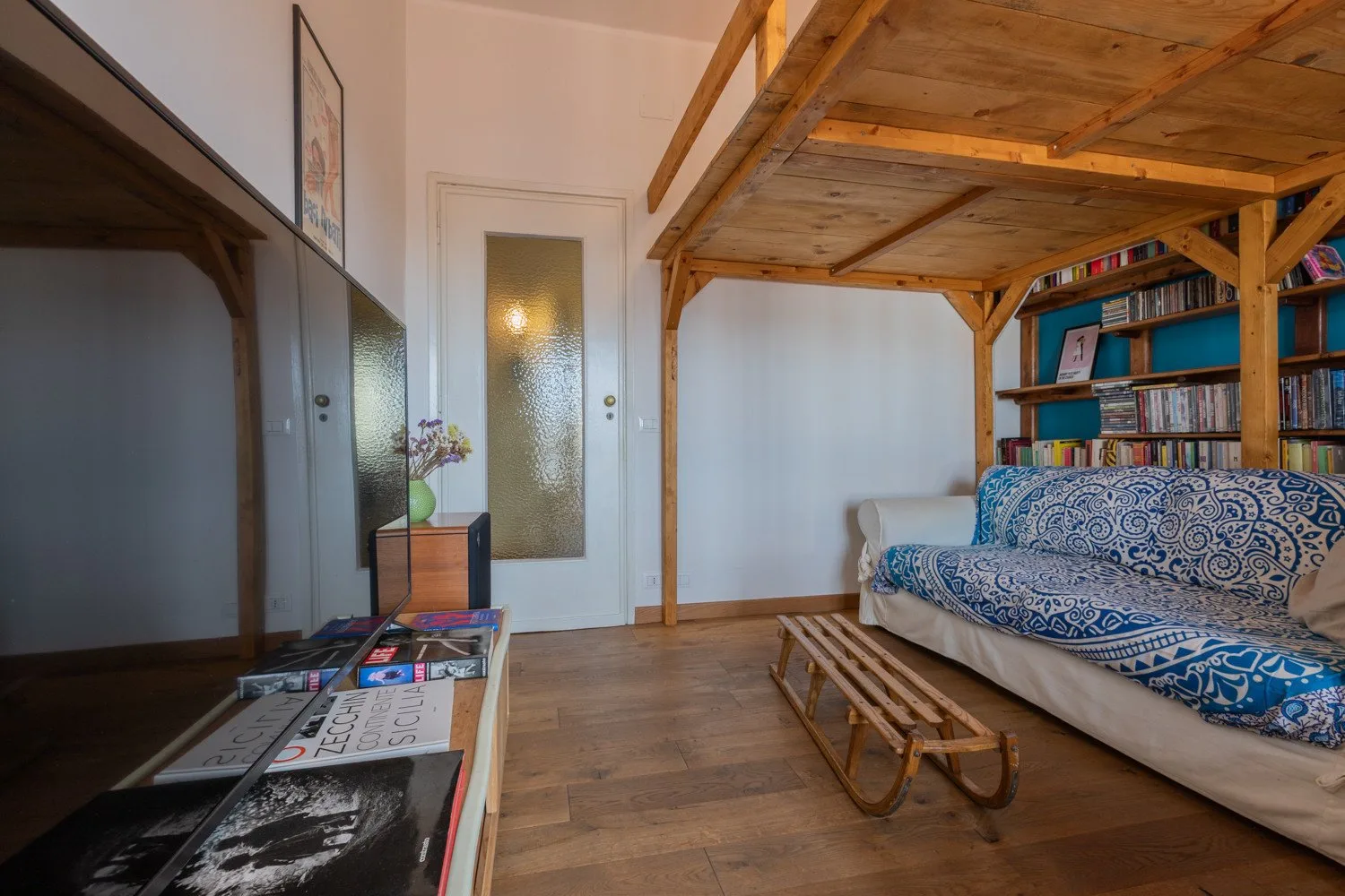 Una sala de estar con un diván bajo un altillo de madera y una estantería.