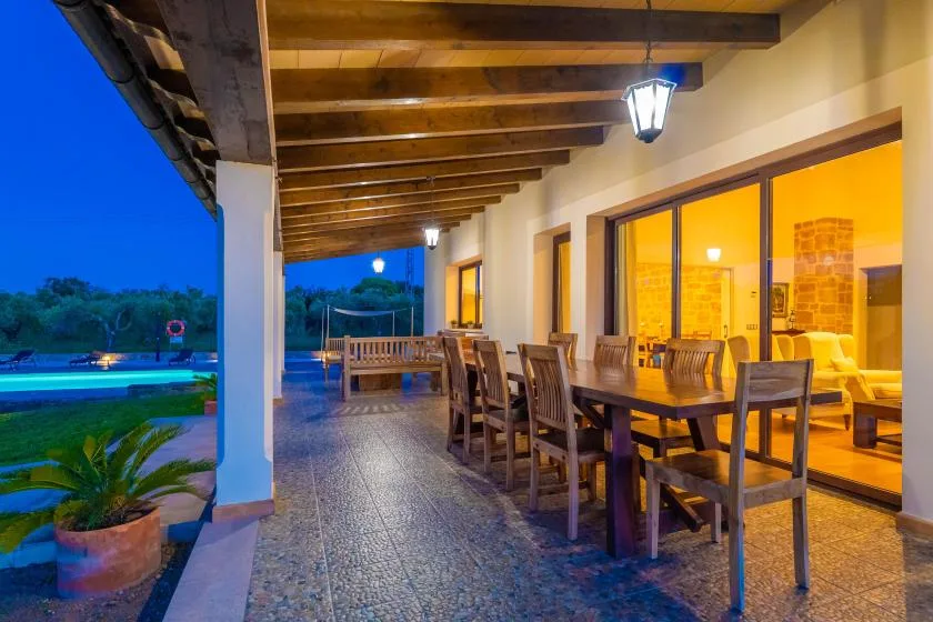 Una zona cubierta de comedor al aire libre con una mesa larga y sillas junto a ventanas iluminadas.