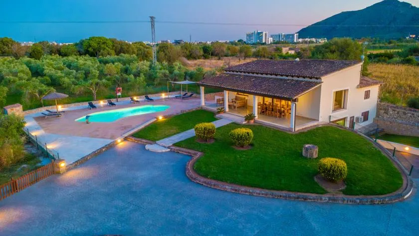 Una casa moderna junto a una piscina iluminada, con jardines y colinas al fondo.