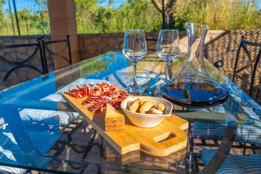 Una mesa de vidrio en el patio sostiene copas, una jarra y una tabla con aperitivos.