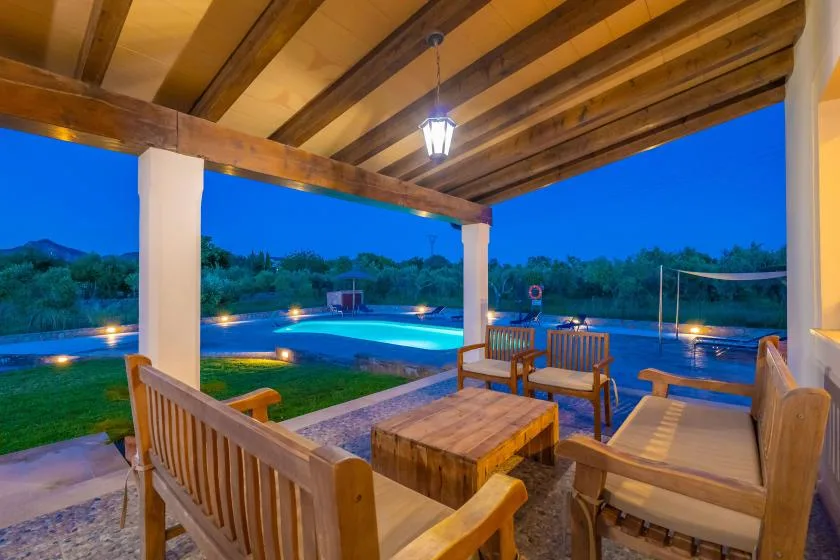 Un patio cubierto con asientos de madera da a una piscina iluminada al anochecer.