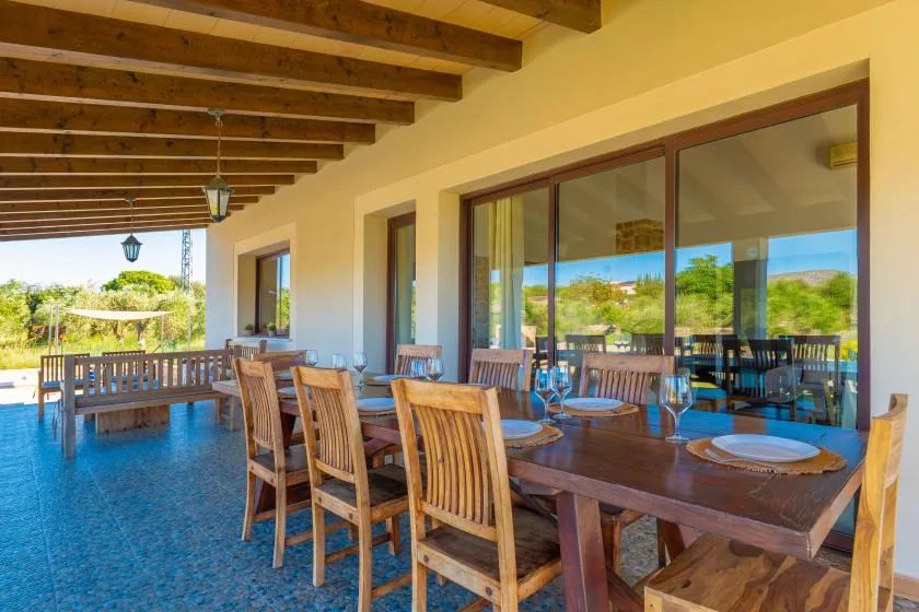 Una zona de comedor exterior cubierta tiene una mesa larga de madera y sillas junto a grandes puertas de vidrio.