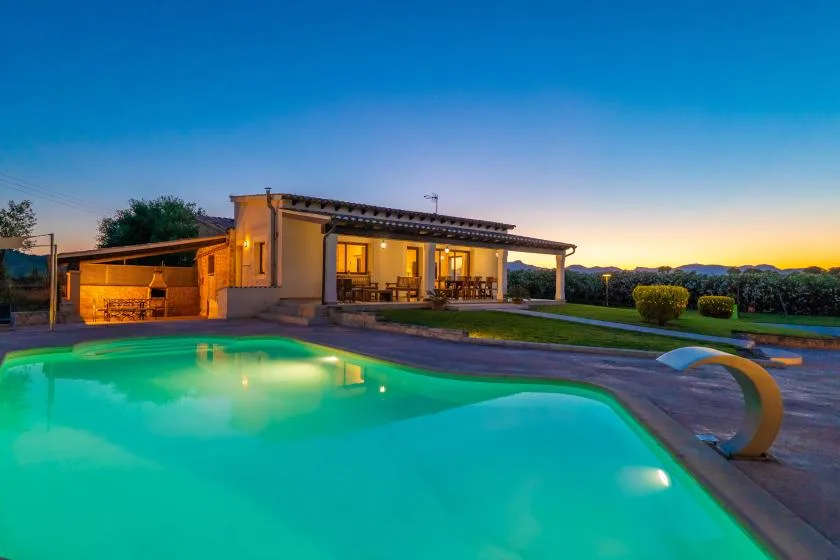 Una piscina iluminada junto a una casa moderna al anochecer.