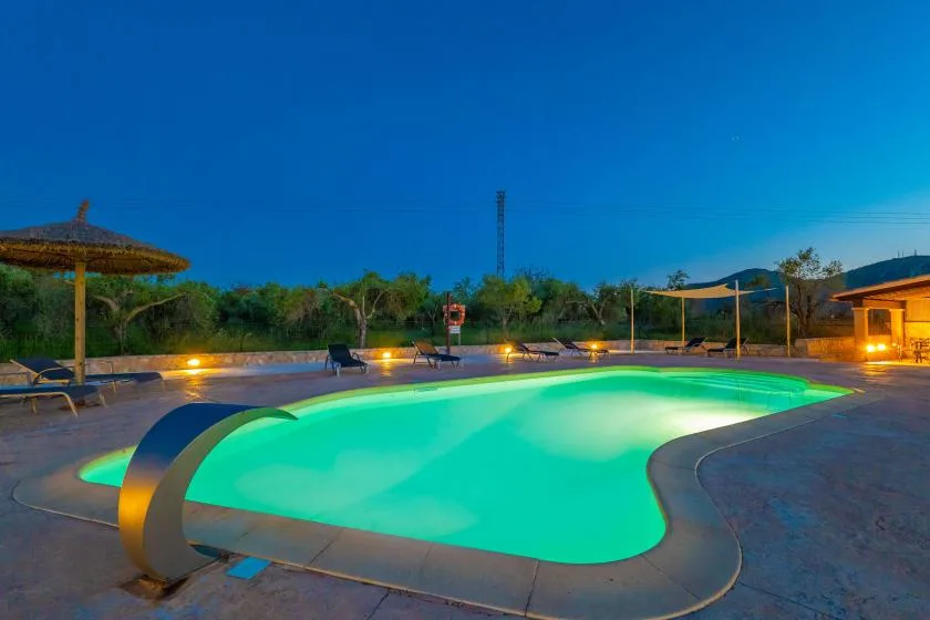 Una piscina iluminada resplandece en tonos turquesa junto a una zona de descanso al anochecer.