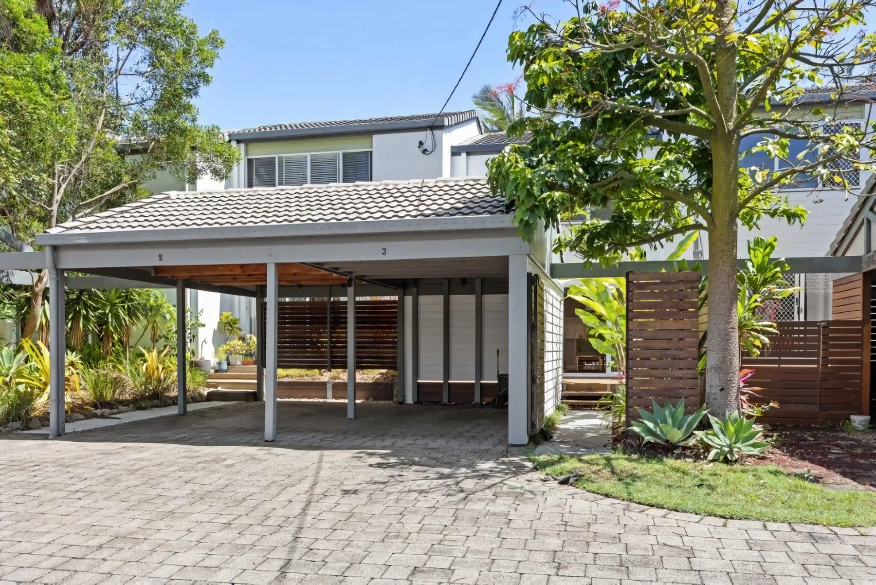 A covered outdoor parking area sits beside a modern house and landscaped garden.