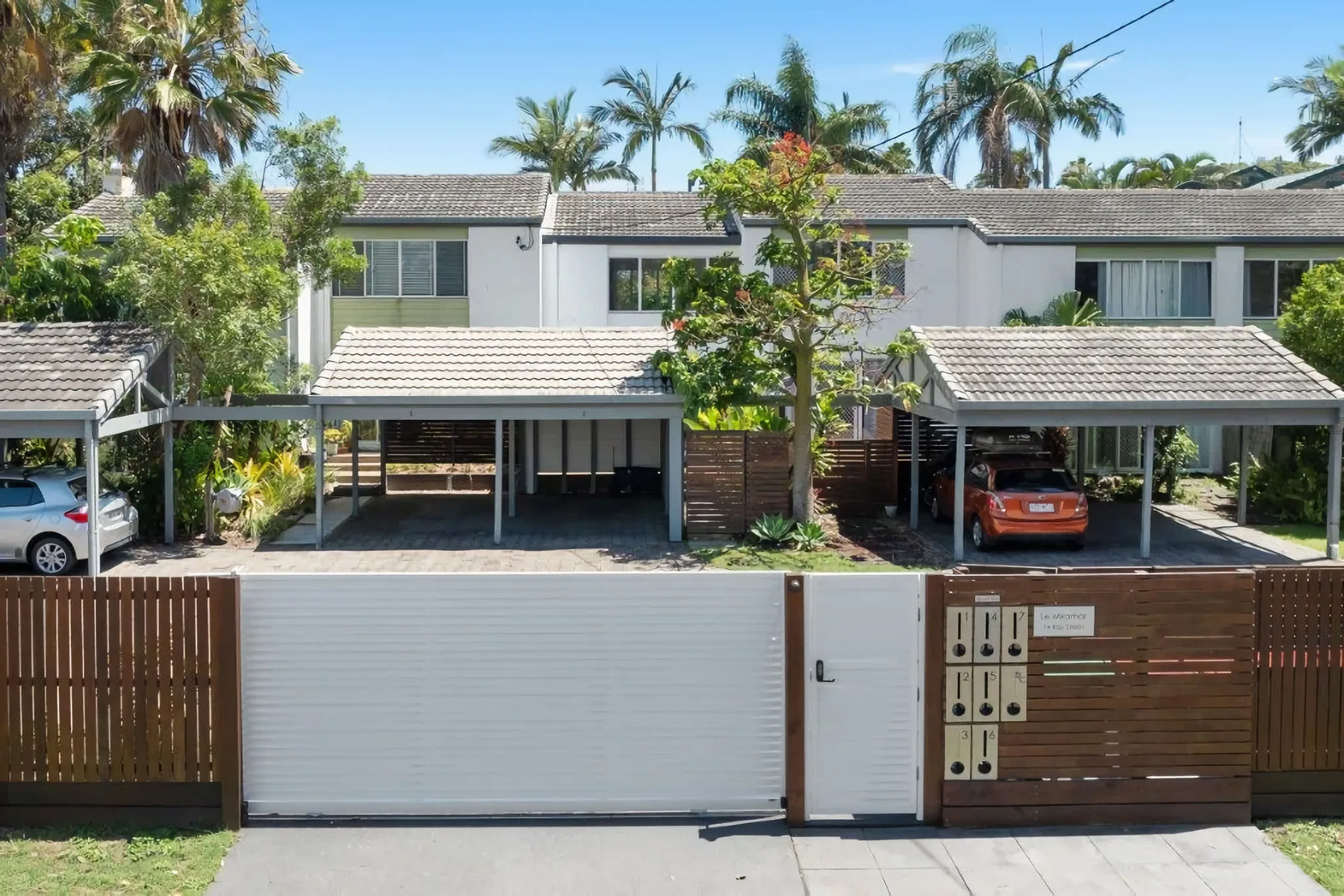A modern two-story house stands behind a gated driveway, with palm trees nearby.