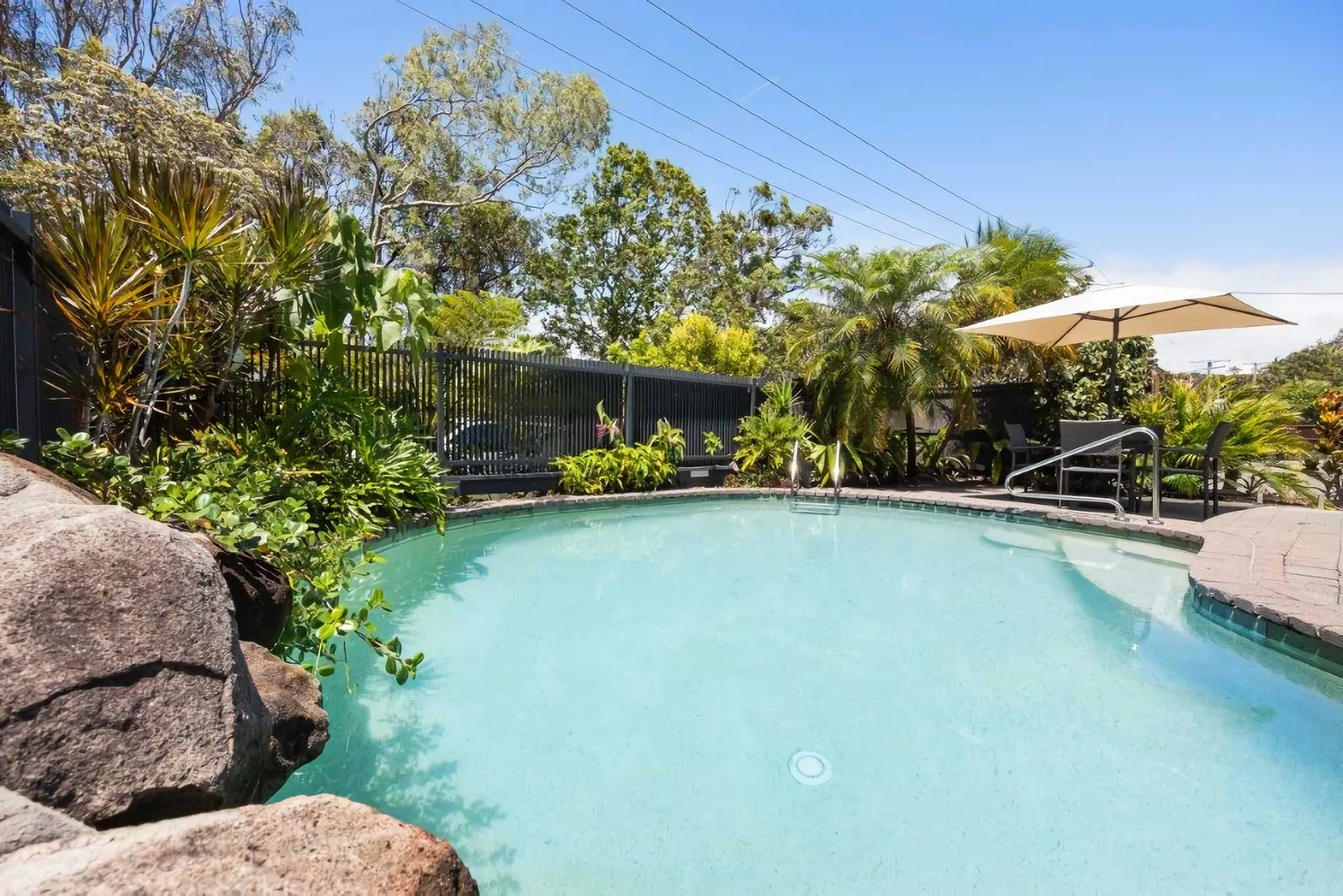 An outdoor swimming pool sits among tropical plants, with an umbrella beside it.