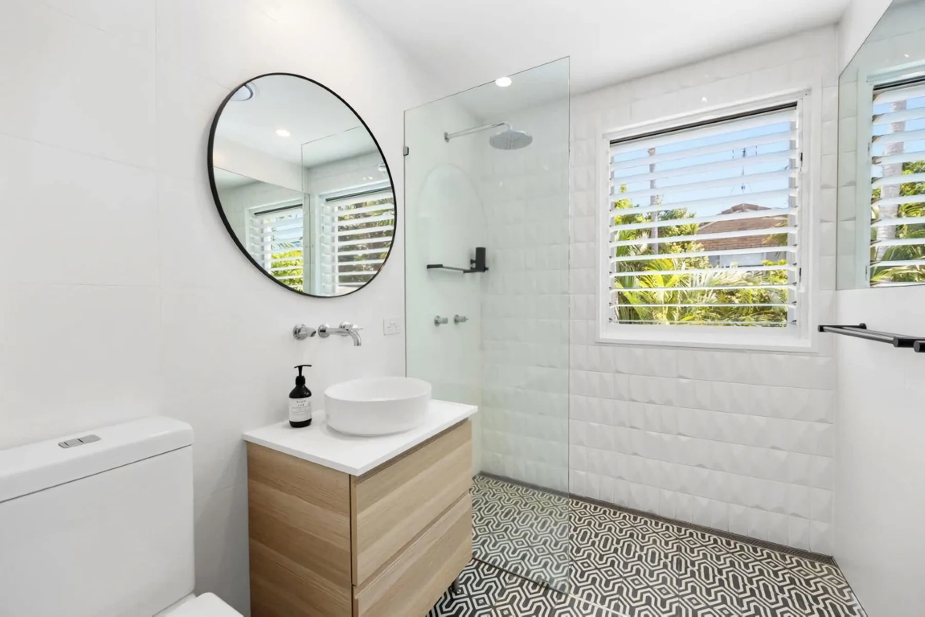 A bright bathroom with a sink vanity, round mirror, and glass shower.