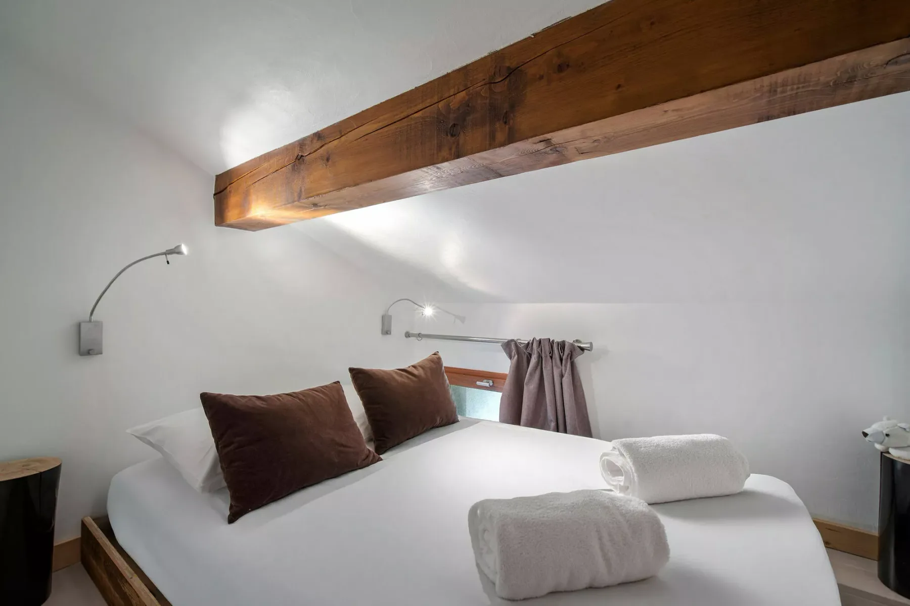 Un dormitorio con una cama bien tendida bajo un techo inclinado y una viga de madera vista.