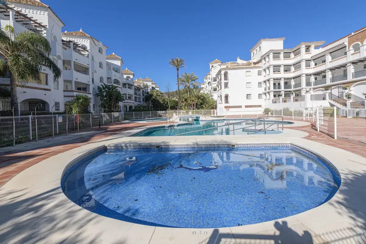 An outdoor swimming pool sits among white apartment buildings and palm trees.