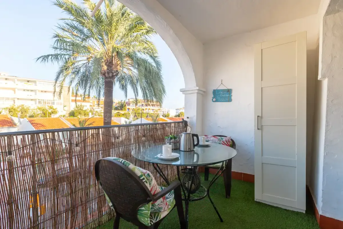 A covered balcony has a small table and seating beside a palm tree.