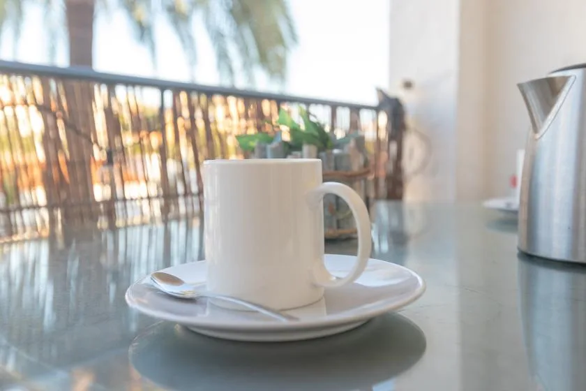 A white mug and saucer sit on a table beside a metal kettle.