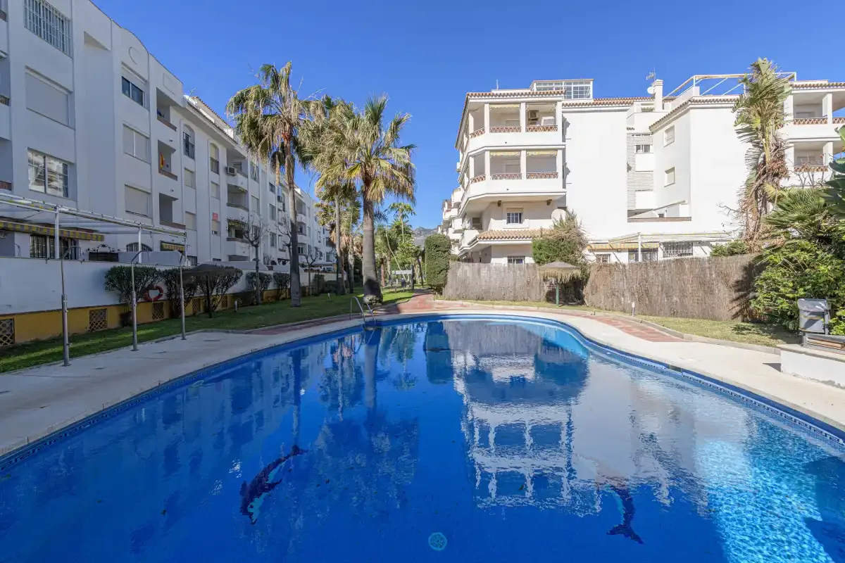 An outdoor swimming pool sits between white apartment buildings and palm trees.