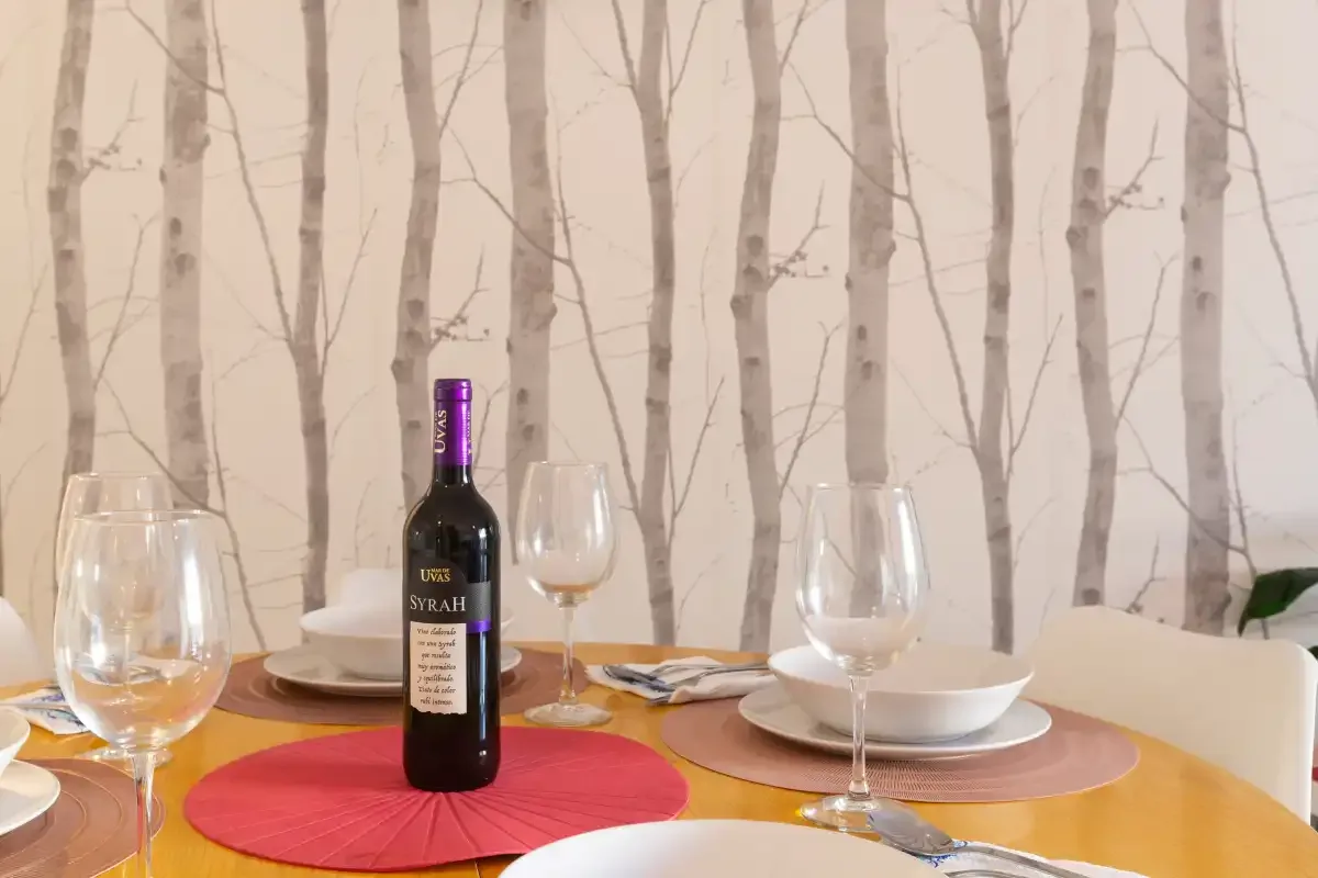 A dining table is set with wine glasses and a bottle in front of a tree-patterned wall.