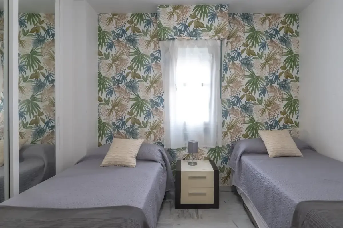 A bedroom with two single beds, leafy wallpaper, and a window.
